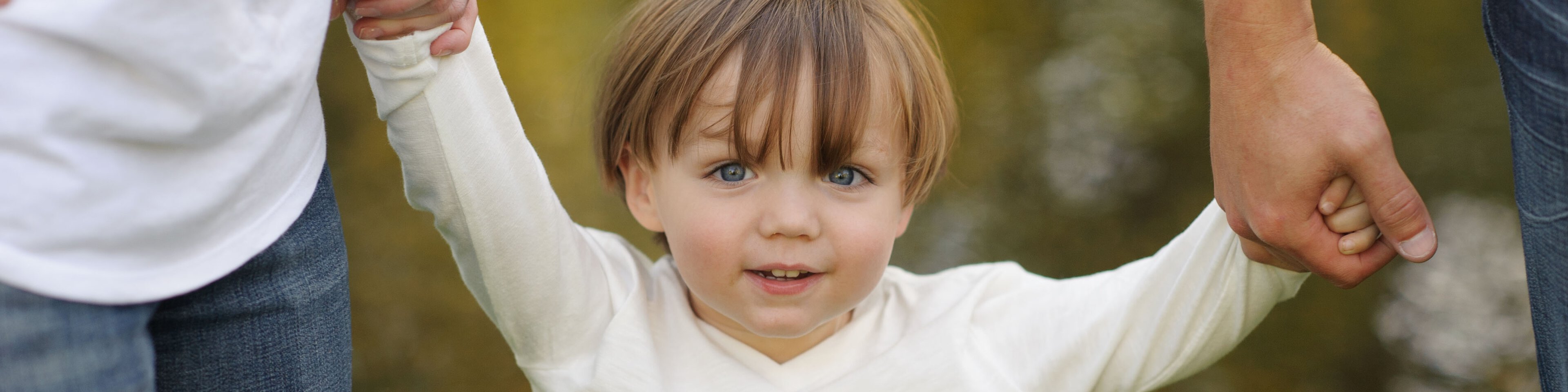 Ein kleiner blonder Junge mit weißem Pullover wird an beiden Händen von seinen Eltern gehalten  | © RealCreation - Getty Images/iStockphoto