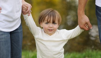 Ein kleiner blonder Junge mit weißem Pullover wird an beiden Händen von seinen Eltern gehalten  | © RealCreation - Getty Images/iStockphoto