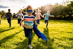 Ein kleiner blonder Junge mit Superman-Verkleidung rennt mit anderen Kindern auf einer Wiese | © StockPlanets - Getty Images