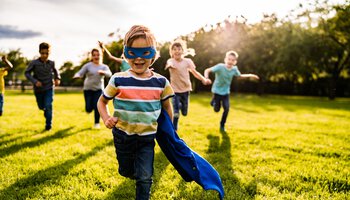 Ein kleiner blonder Junge mit Superman-Verkleidung rennt mit anderen Kindern auf einer Wiese | © StockPlanets - Getty Images